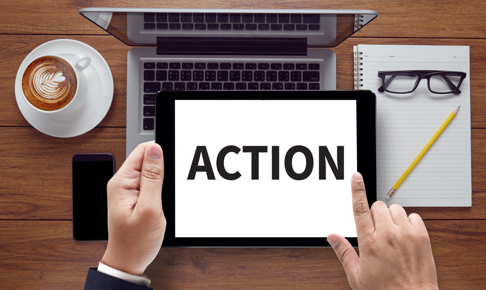 A man is holding a sign with the word, Action, on a tablet.  A laptop is in the background.