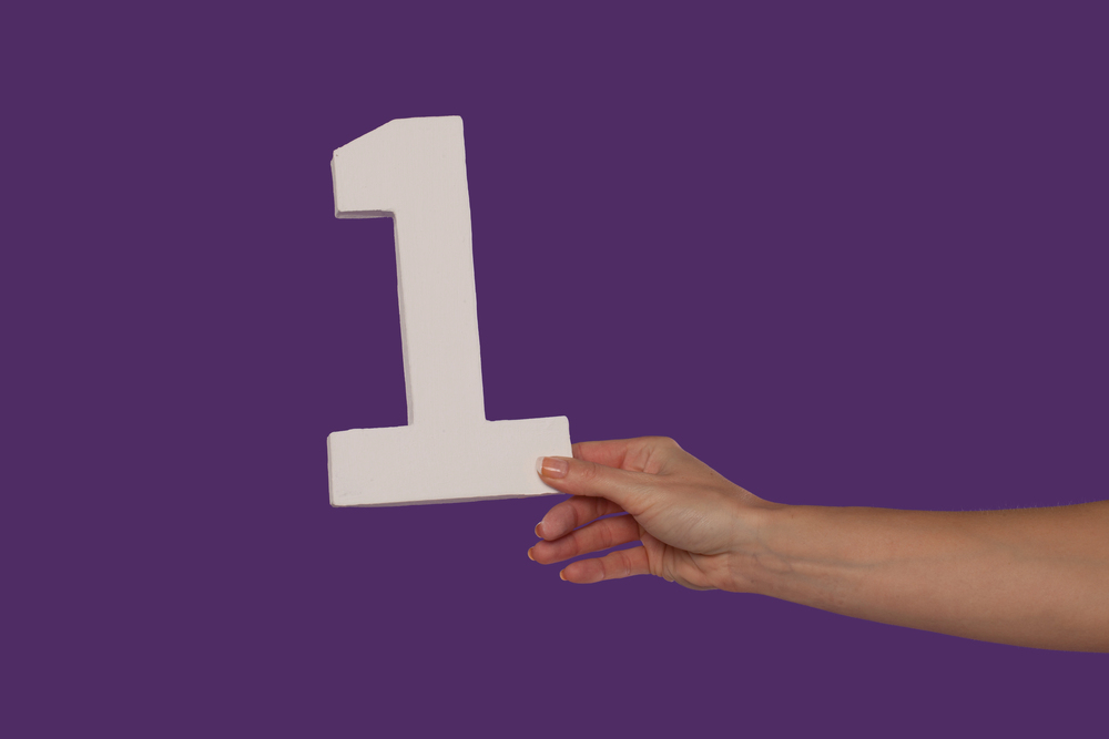 A person is holding a number 1 sign against a purple background. 