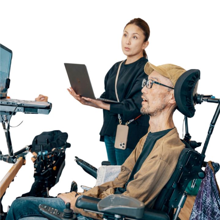 A man in a power wheelchair is using technology to communicate while a lady holding a laptop looks on.