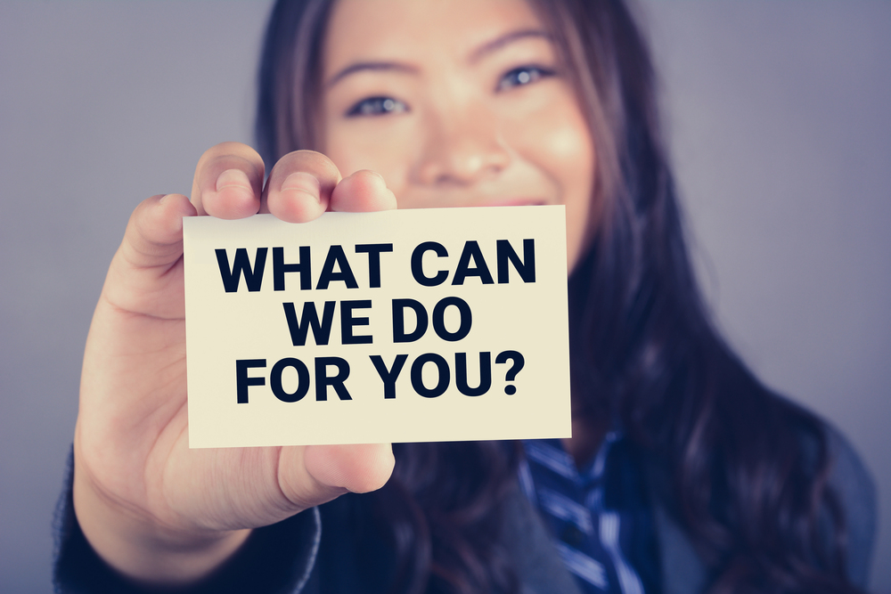 A lady is holding a sign that asks the question, "How can we help you?"