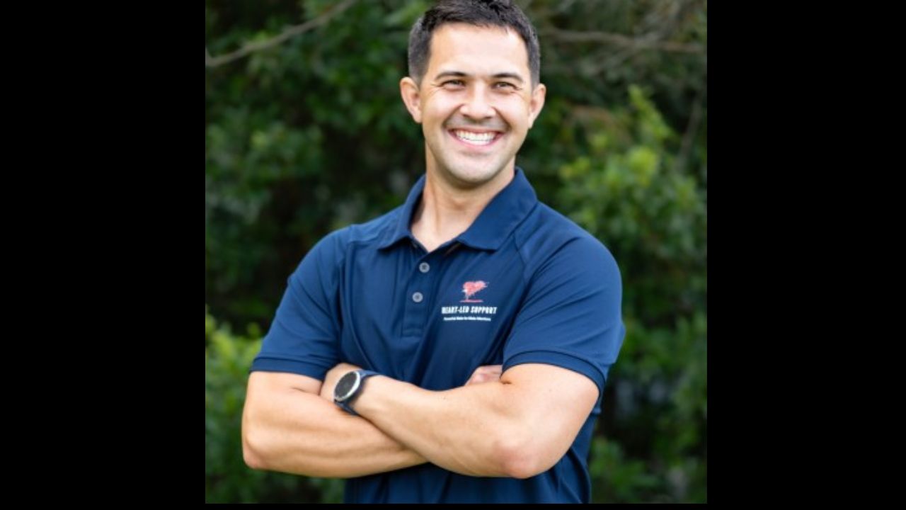 Joseph Lee is standing with his arms crossed against a wall of trees.