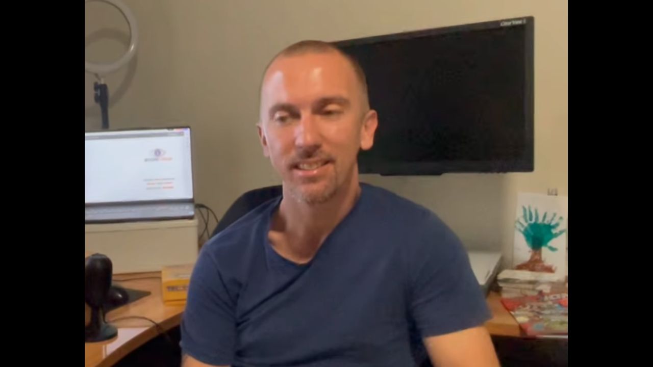 Peter Archer is sitting in an office with computer screens behind him.