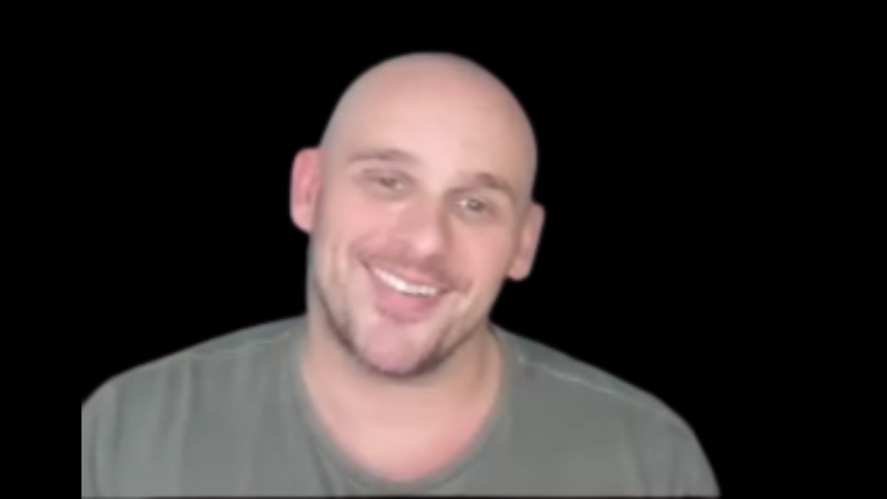 Tommy Trout is wearing a grey t-shirt and is leaning against a black background.