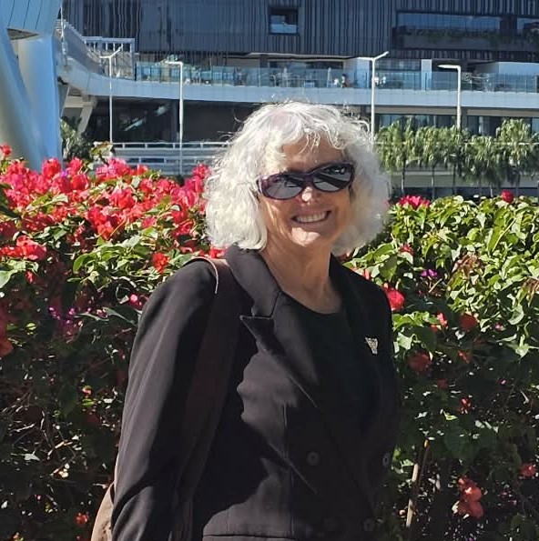 Narelle is wearing a black jacket and is standing in front of a hedge.